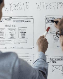 Man holding a red-capped whiteboard marker and pointing it at a whiteboard with website wireframe mockups
