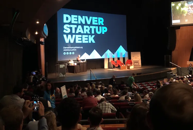 Indoor stage featuring a large projector screen with the words Denver Startup Week behind a stage with three panelist chairs.