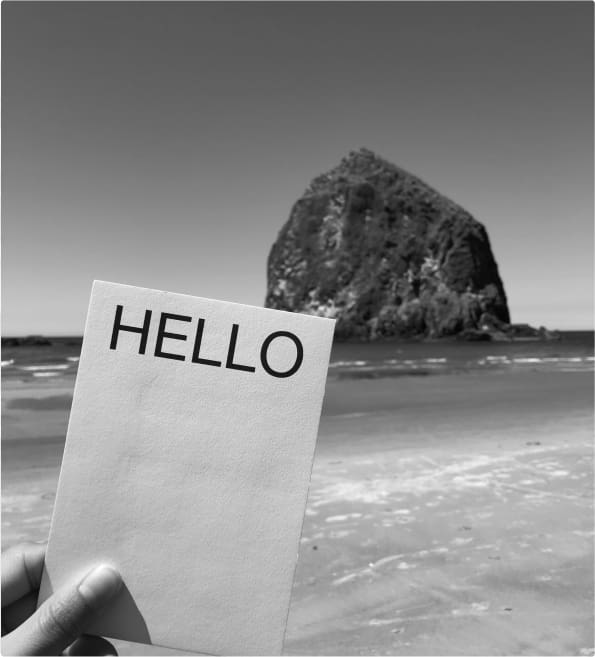 A white postcard with the word "hello" printed prominently in black held in front of a beach scene with a large rock in the foreground.