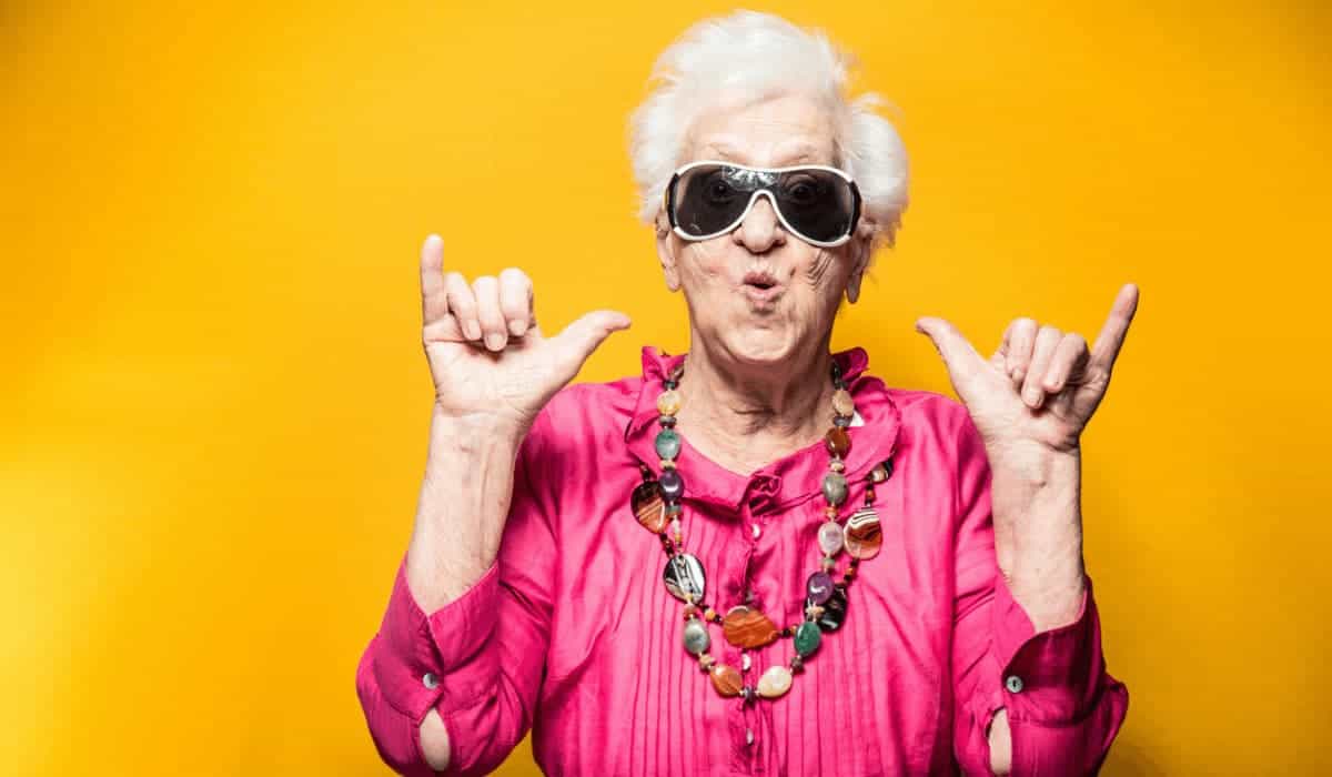 an older woman with short gray hair wearing sunglasses and making a humorous face while holding up "hang loose" hand gestures with both hands, set against a bright yellow background